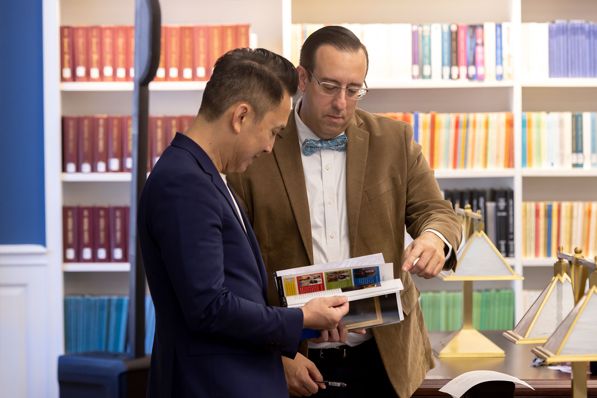 Librarian and patron look through the pages of a book inside Bridwell Library, with shelves of books on the wall behind them.